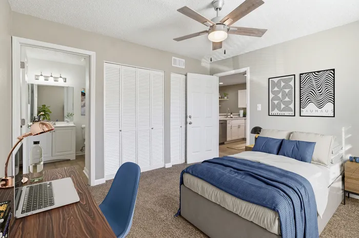 Charming bedroom with cozy bed, modern ceiling fan, and a workspace featuring a sleek laptop, leading to a stylish bathroom and open kitchen area.