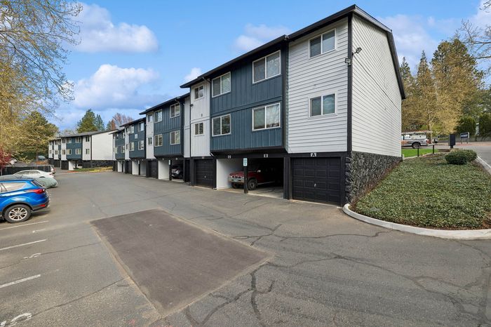 Charming apartment complex featuring modern exteriors with clean lines and attached garages, set against a backdrop of lush greenery and a clear blue sky.