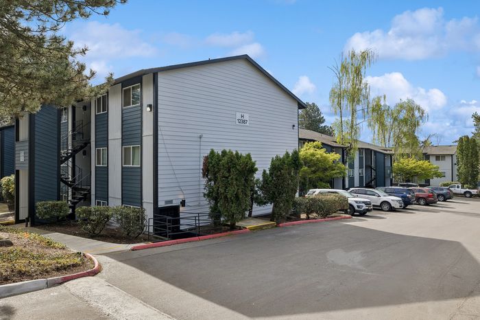Inviting apartment complex exterior with modern blue and white facade, lush landscaping, and ample parking under a bright blue sky.