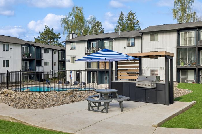 Vibrant apartment complex showcasing a stylish outdoor area with a modern barbecue grill, picnic table under a chic striped umbrella, and a refreshing swimming pool enclosed by a safety fence.