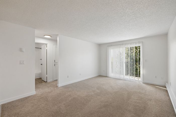 Bright and spacious living area with plush carpet and sliding glass doors leading to a lush outdoor view.