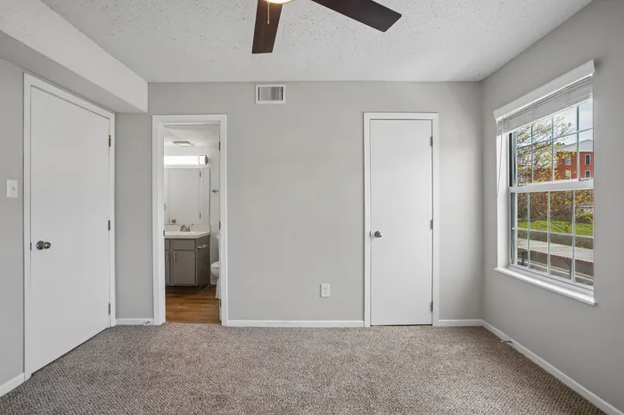 Bright bedroom with plush carpet, modern ceiling fan, and a window showcasing a scenic view, connected to a sleek, stylish bathroom.