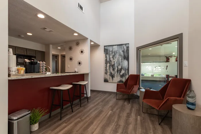 Chic apartment interior featuring a sleek breakfast bar with high stools, modern artwork, and cozy seating area with red velvet chairs.