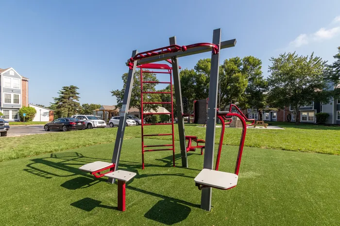 Modern outdoor fitness area with sleek red exercise equipment set against a backdrop of lush green grass and a scenic residential neighborhood.