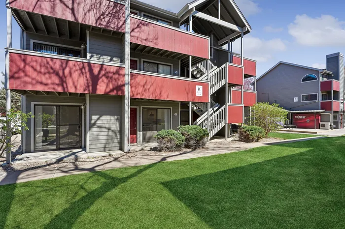Charming multi-level apartment building with vibrant red accents, surrounded by lush green landscaping and a clear blue sky.