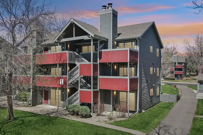 Charming three-story apartment complex with vibrant red balconies and lush green surroundings, set against a colorful sunset sky.