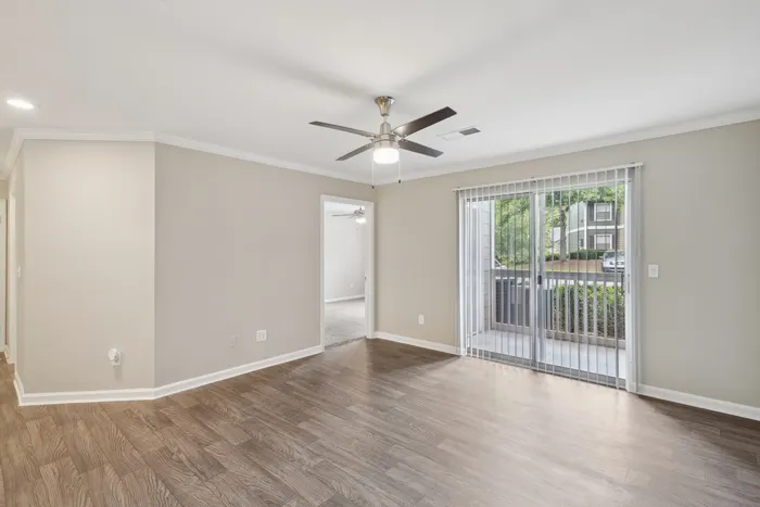 Bright and airy apartment living room with elegant wood flooring, ceiling fan, and large sliding glass door leading to a serene outdoor view.