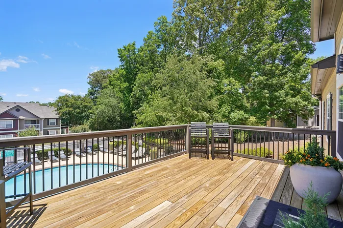 Charming apartment balcony with wooden deck overlooking a refreshing pool and lush green trees, perfect for relaxation.