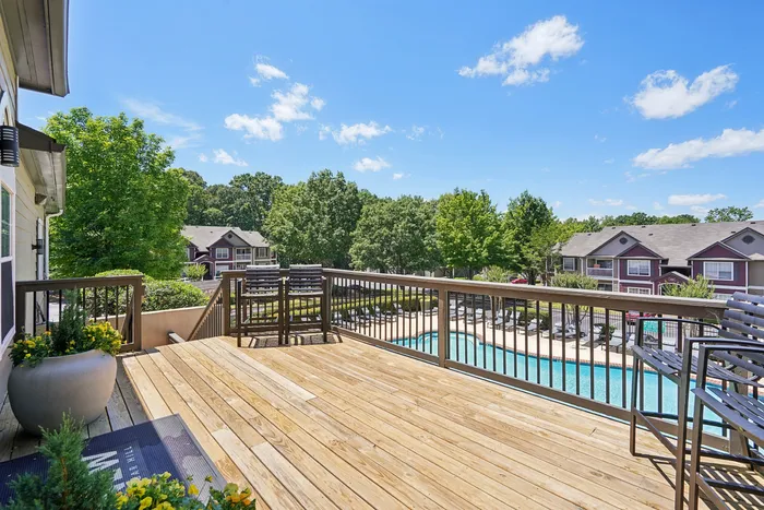 Inviting wooden deck with lush greenery, overlooking a sparkling community pool and charming residential buildings under a bright, blue sky.