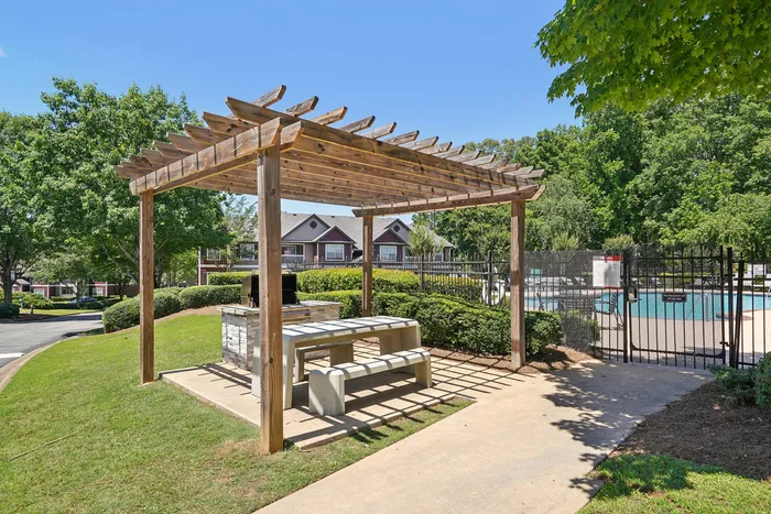 Charming outdoor space featuring a wooden pergola with picnic benches, perfect for gatherings, set against a backdrop of lush greenery and a sparkling swimming pool.