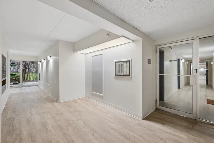 Bright and inviting apartment entryway featuring sleek wood flooring, modern lighting, and a view of lush greenery through the glass door.