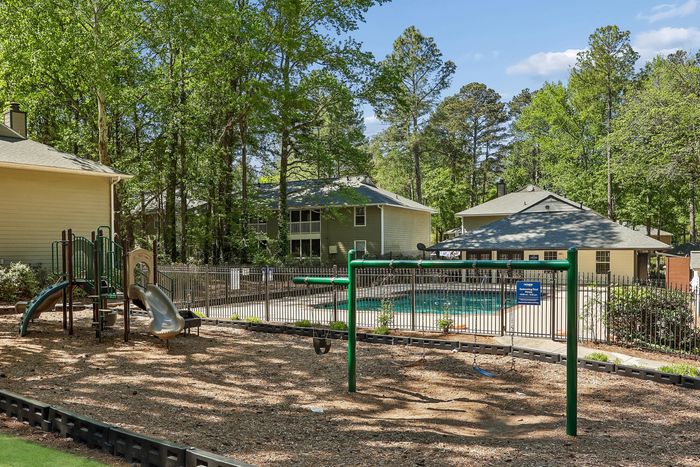 Charming apartment complex with inviting playground and sparkling pool, nestled among lush green trees.