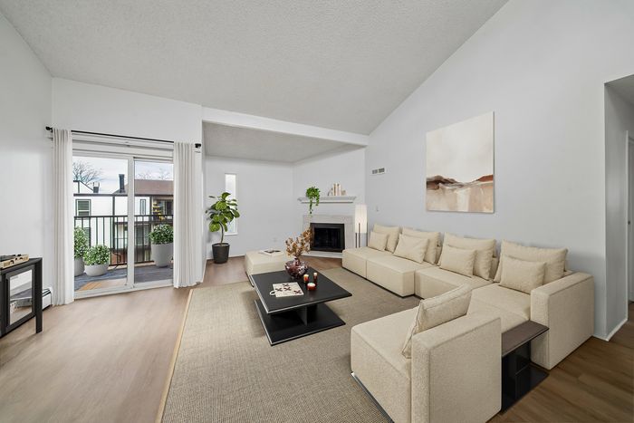Bright and spacious living room featuring a cozy beige sectional sofa, a modern coffee table, and a decorative fireplace, with large windows leading to a balcony with potted plants.