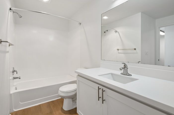 Modern white bathroom featuring a sleek vanity with a wide mirror, elegant fixtures, bathtub, and natural wood flooring.