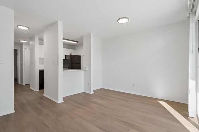 Modern open-concept living area with sleek hardwood floors and a view into a minimalist kitchen featuring white cabinetry and stainless steel appliances.