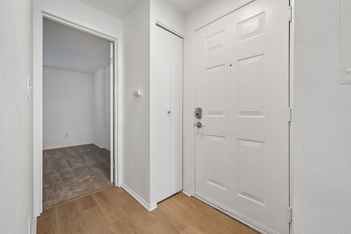 Inviting apartment entryway with elegant wood flooring and fresh white walls, leading into a cozy carpeted room.