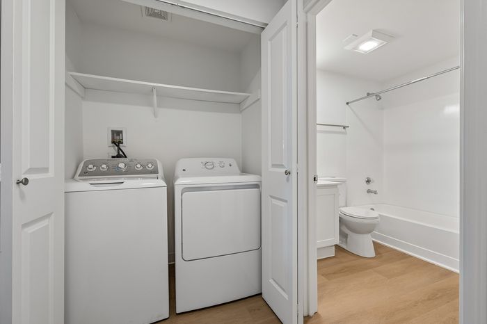 Modern apartment laundry and bathroom area featuring in-unit washer and dryer next to a sleek, white bathroom with a bathtub and wood-style flooring.