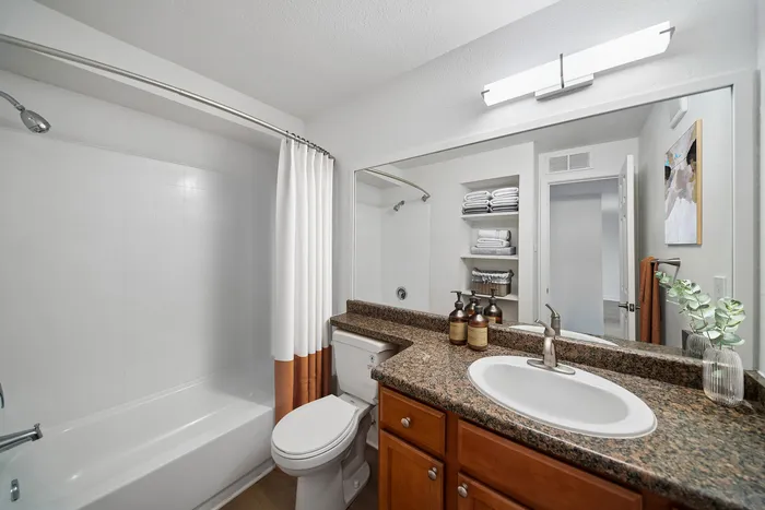Modern bathroom with granite countertop, elegant fixtures, and abundant natural light, featuring a clean white tub and neatly organized open shelving.