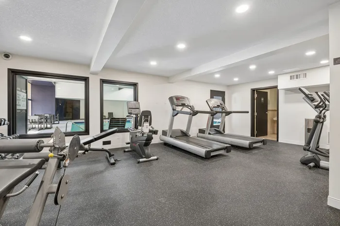 Modern apartment gym with treadmills, exercise bike, and weight machine, featuring bright lighting and pool view.