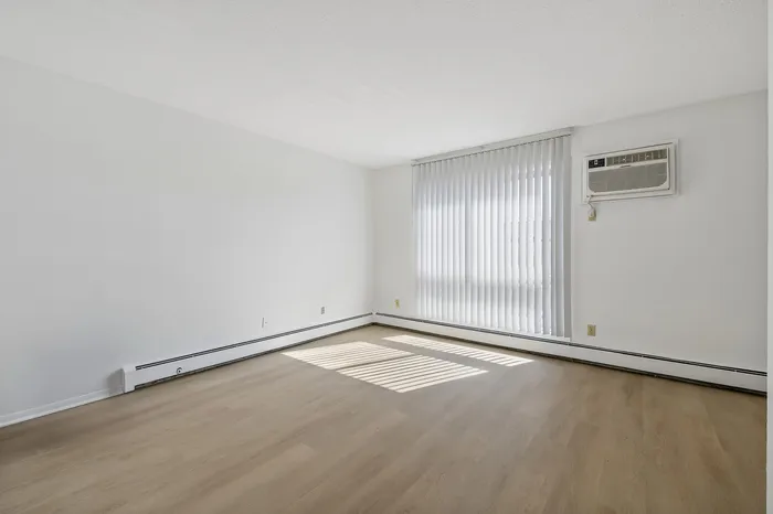 Bright and inviting living room with sleek wooden floors, large window with vertical blinds, and ample natural light perfect for cozy living.