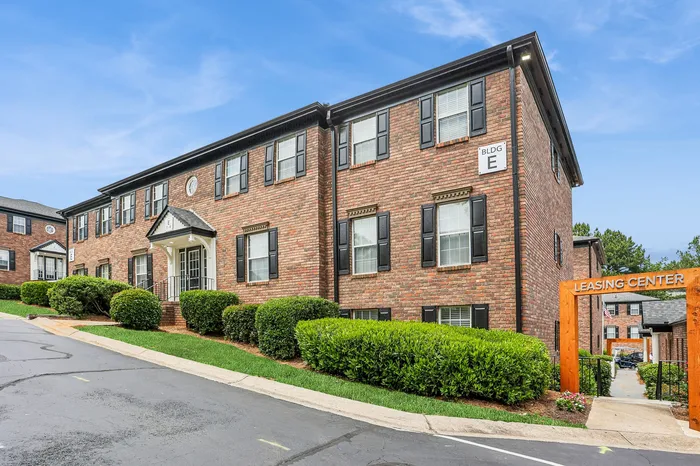 Charming brick apartment building with manicured hedges and a welcoming leasing center entrance under a clear blue sky.