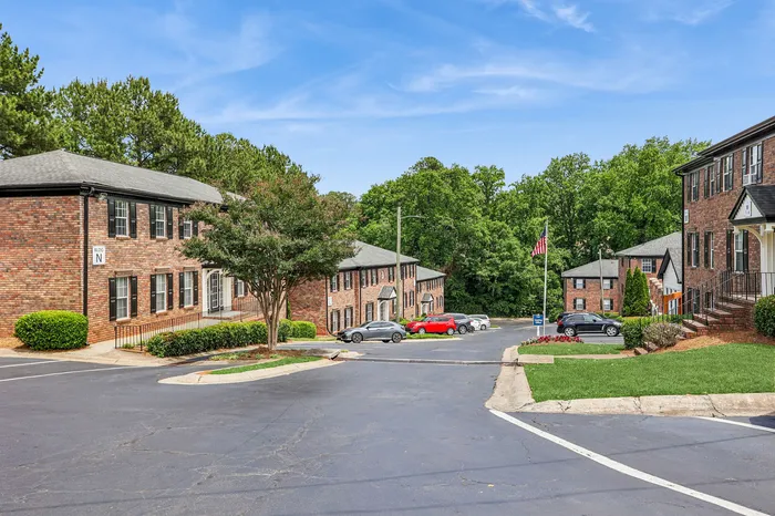 Charming suburban apartment community with brick exteriors, lush greenery, and ample parking spaces against a clear blue sky.