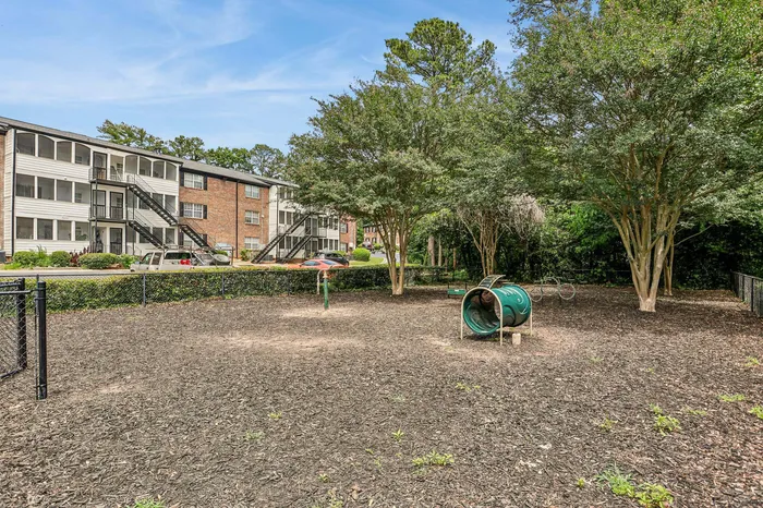 Charming apartment complex with well-maintained exteriors and inviting landscaping, featuring a fenced playground area surrounded by lush trees.