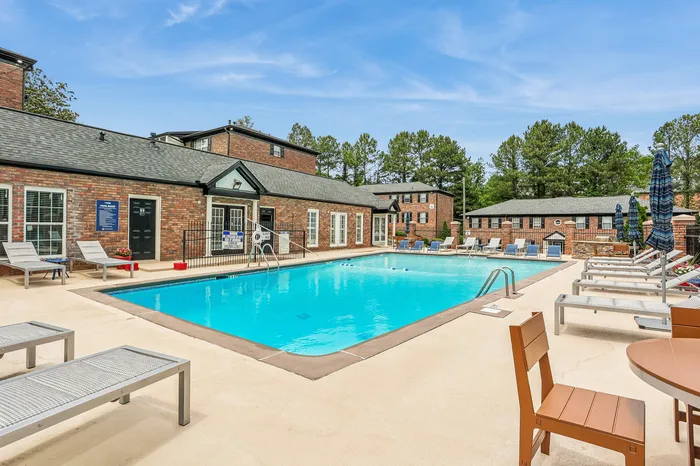 Inviting outdoor pool area with stylish lounge chairs and brick clubhouse, surrounded by lush greenery and clear blue skies.