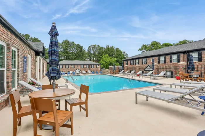 Inviting apartment complex pool area with modern lounge chairs, elegant umbrellas, and vibrant greenery under a bright blue sky.