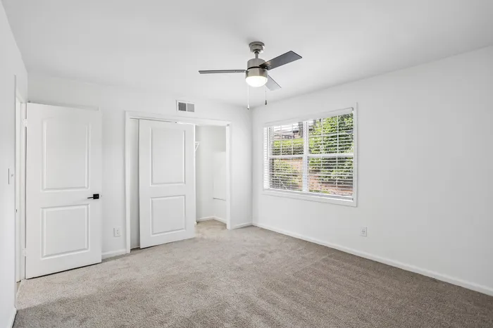 Bright and modern bedroom with plush carpet, sleek ceiling fan, and ample natural light from large window overlooking greenery.