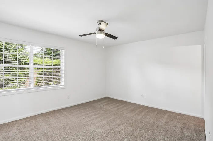Bright, airy bedroom with plush carpeting, ceiling fan, and large window overlooking lush greenery.