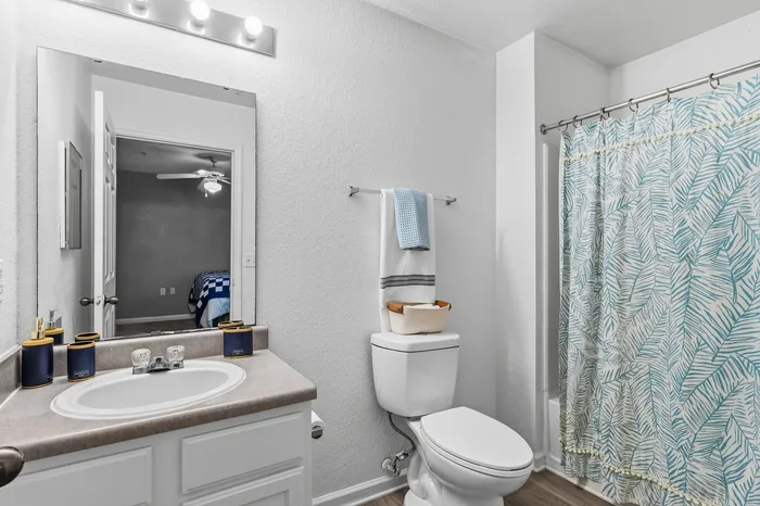 Elegant bathroom with a modern vanity, chic lighting, and tropical-themed shower curtain, featuring a serene view into a cozy bedroom.