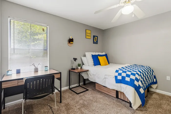 Inviting bedroom with cozy bed, vibrant blue and yellow accents, stylish workspace, and natural light streaming through a large window.