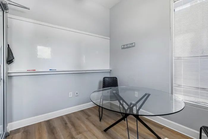 Modern private office space featuring a sleek glass round table, comfortable chair, large whiteboard, and abundant natural light through blinds.