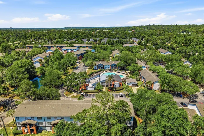 Vibrant aerial view of a serene apartment complex surrounded by lush greenery, featuring a central swimming pool and charming residential buildings.
