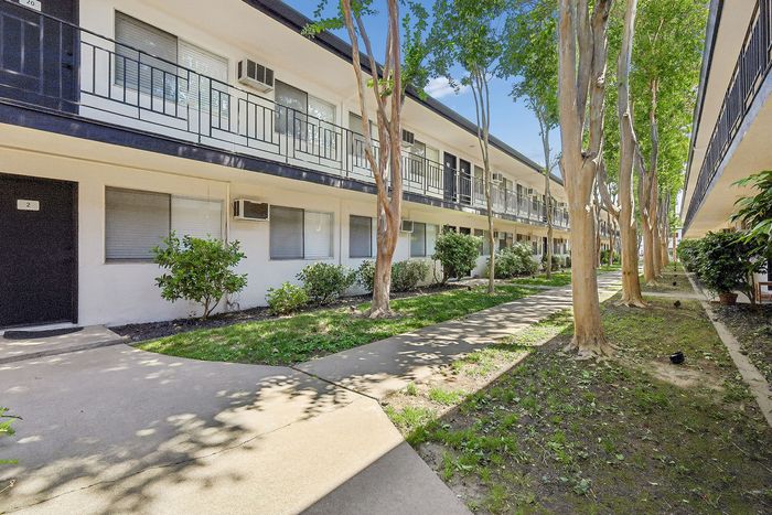 Charming apartment complex with two-level walkways, surrounded by lush greenery and tall trees, offering a serene living environment.