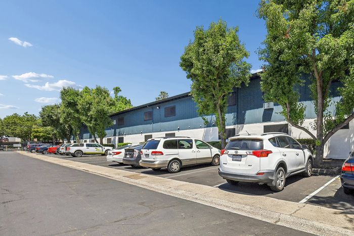 Vibrant exterior of an apartment building with ample parking, lush trees, and clear blue skies, perfect for a sunny lifestyle.