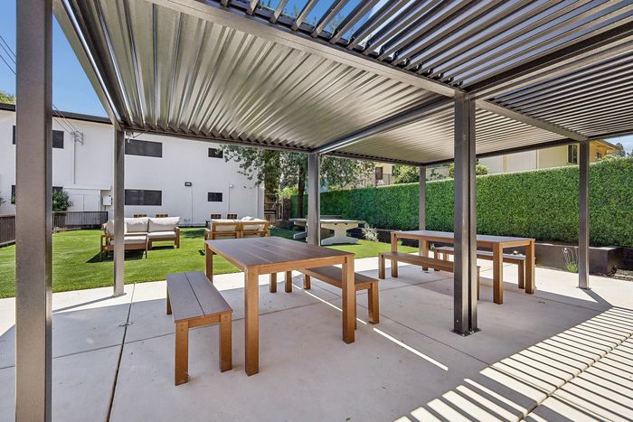 Charming outdoor patio area with wooden picnic tables under a modern pergola, surrounded by lush greenery and comfortable seating, perfect for gatherings.