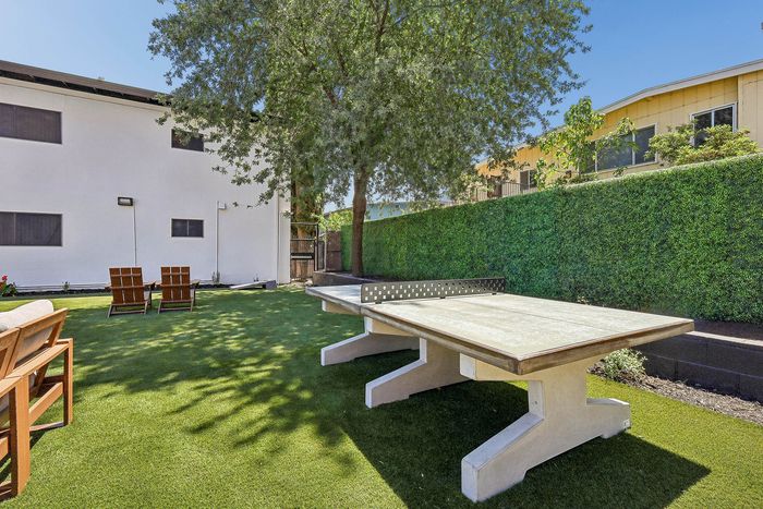 Charming outdoor space featuring a ping pong table, lush green lawn, and cozy wooden chairs under the shade of a large tree.