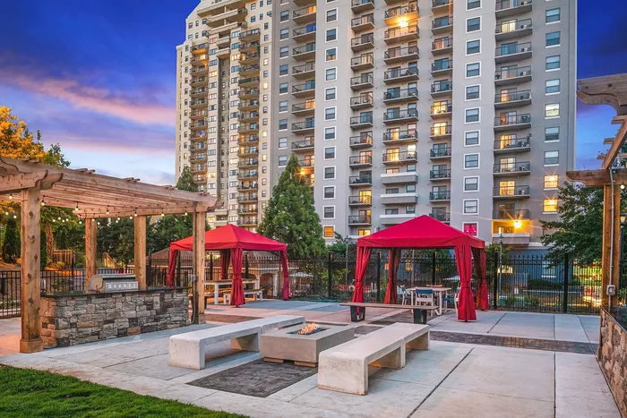 Vibrant apartment complex featuring a modern outdoor lounge area with red cabanas, stylish pergola, and ambient lighting, set against a sunset skyline.