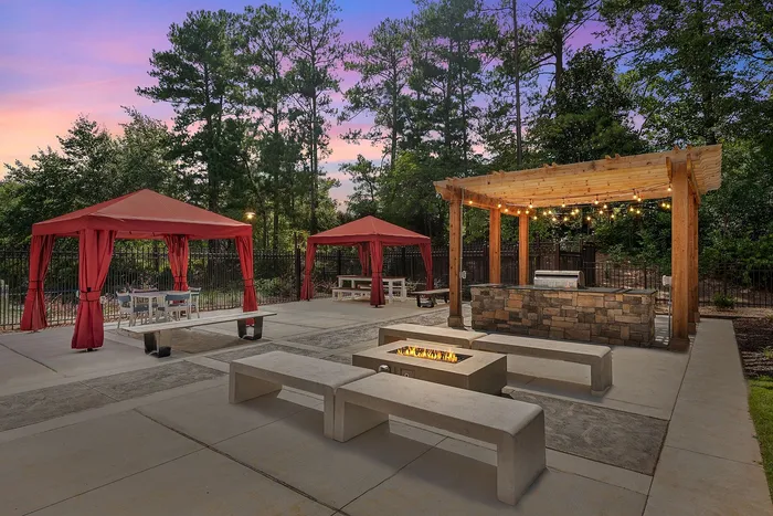 Charming outdoor patio with stone grill station and cozy fire pit, featuring red canopy seating and surrounded by lush greenery at sunset.