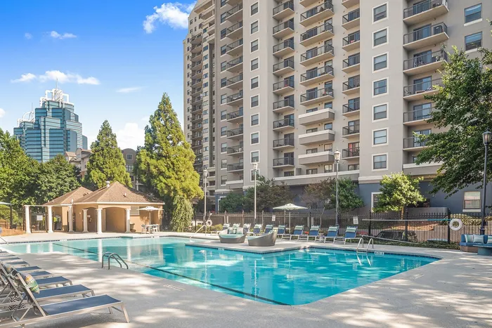 Modern apartment complex with a luxurious swimming pool, surrounded by lush greenery and lounge chairs, under a clear blue sky.