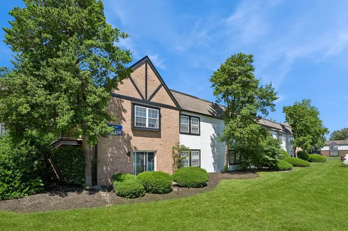 Charming brick apartment building with Tudor-style accents, surrounded by lush greenery and vibrant landscaping under a clear blue sky.