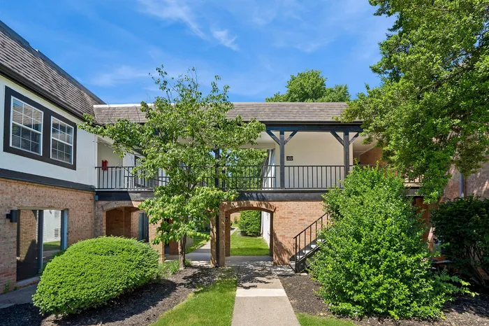 Charming brick apartment exterior with lush greenery and a serene upper walkway underneath a sunny blue sky.