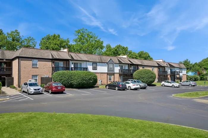 Charming brick apartment building with balconies set against a backdrop of lush greenery, featuring a well-maintained parking area under a bright blue sky.