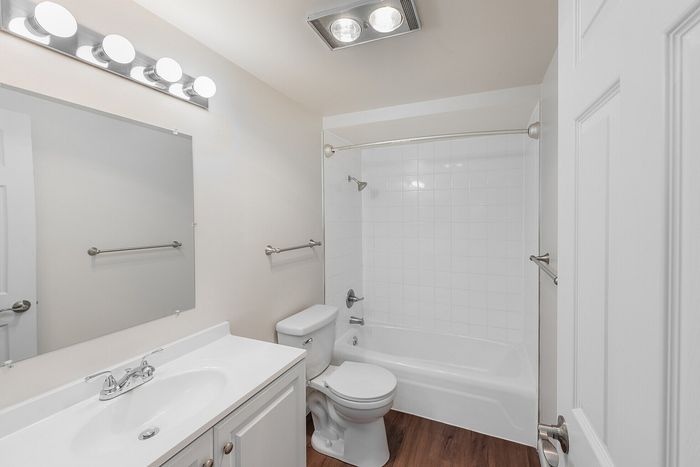 Modern bathroom featuring a sleek white vanity, spacious mirror, and elegant wood flooring. Bright lighting emphasizes the clean, contemporary design.