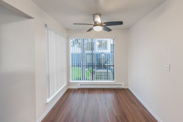 Bright, open room with sleek hardwood flooring and vertical blinds revealing lush outdoor views, complemented by a modern ceiling fan.