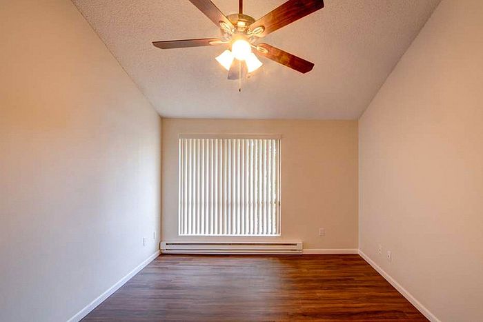 Bright and airy room featuring elegant wood floors, a ceiling fan with warm lighting, and large window with vertical blinds for ample natural light.