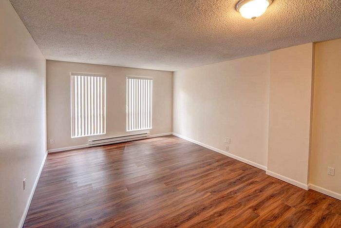 Spacious living room with warm hardwood flooring and natural light streaming through large windows with vertical blinds.