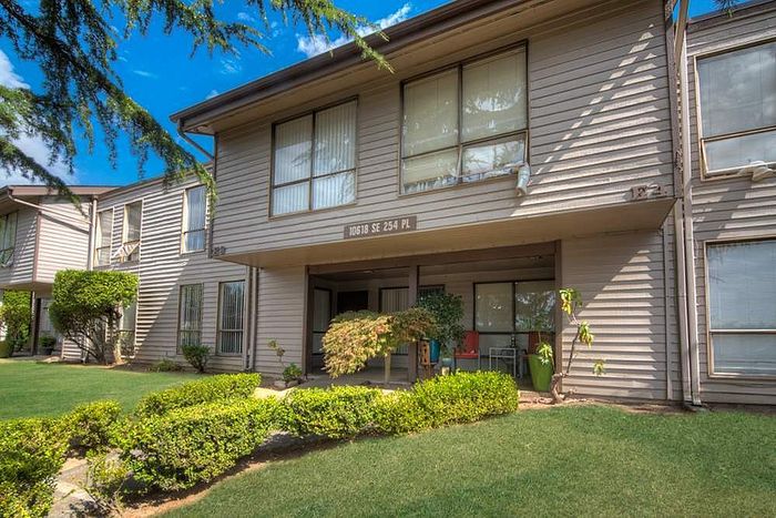 Charming apartment complex with well-kept lawn, large windows, and inviting entryway, surrounded by lush greenery under a clear blue sky.